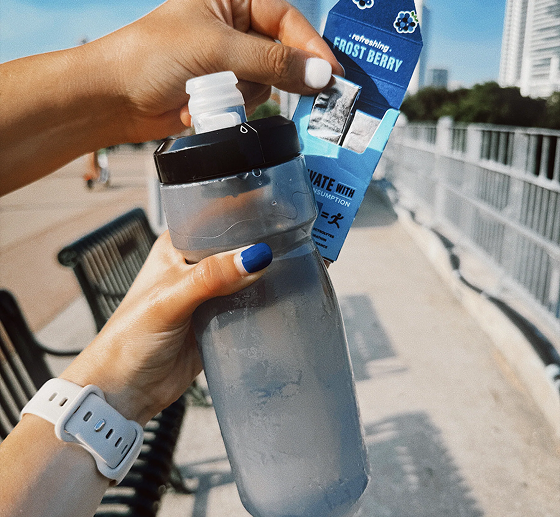 picture of two peoples hands, one holding a water bottle and another holding an pen pack of rev frost berry electrolyte gum, city scape in background
